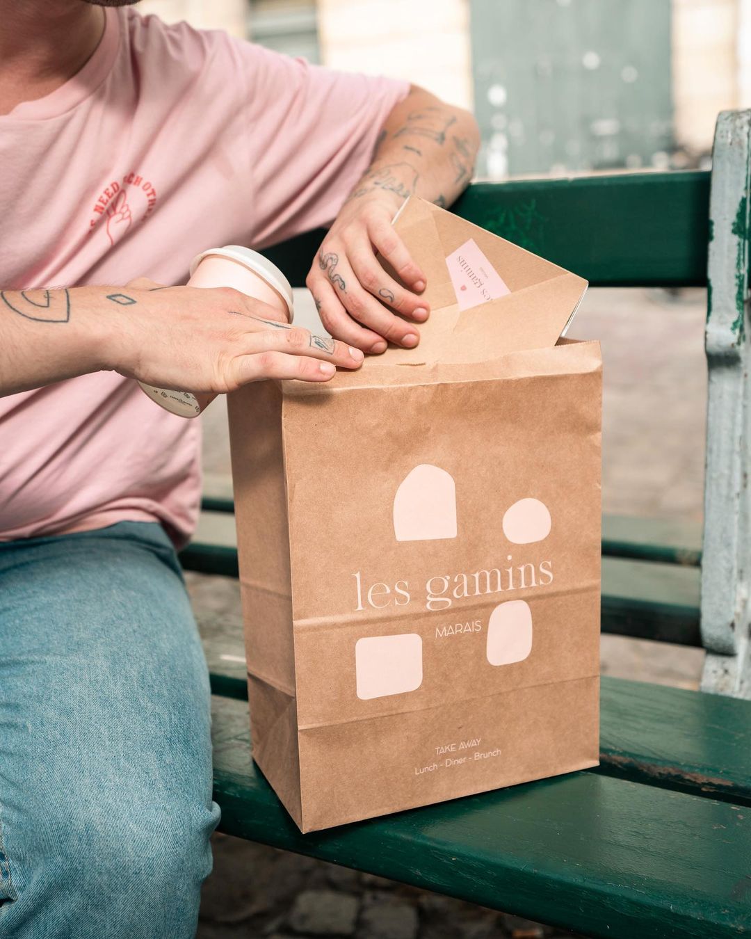 Person sitting on a bench placing a takeaway box into a Les Gamins block bottom paper bag, while holding a coffee cup